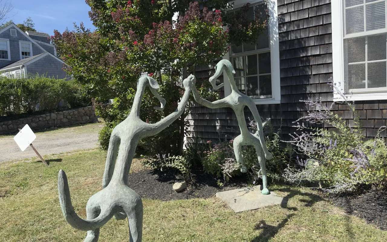 A green-patina sculpture of two long-limbed figures dancing hand in hand in a front garden beside a shingled house in Vineyard Haven