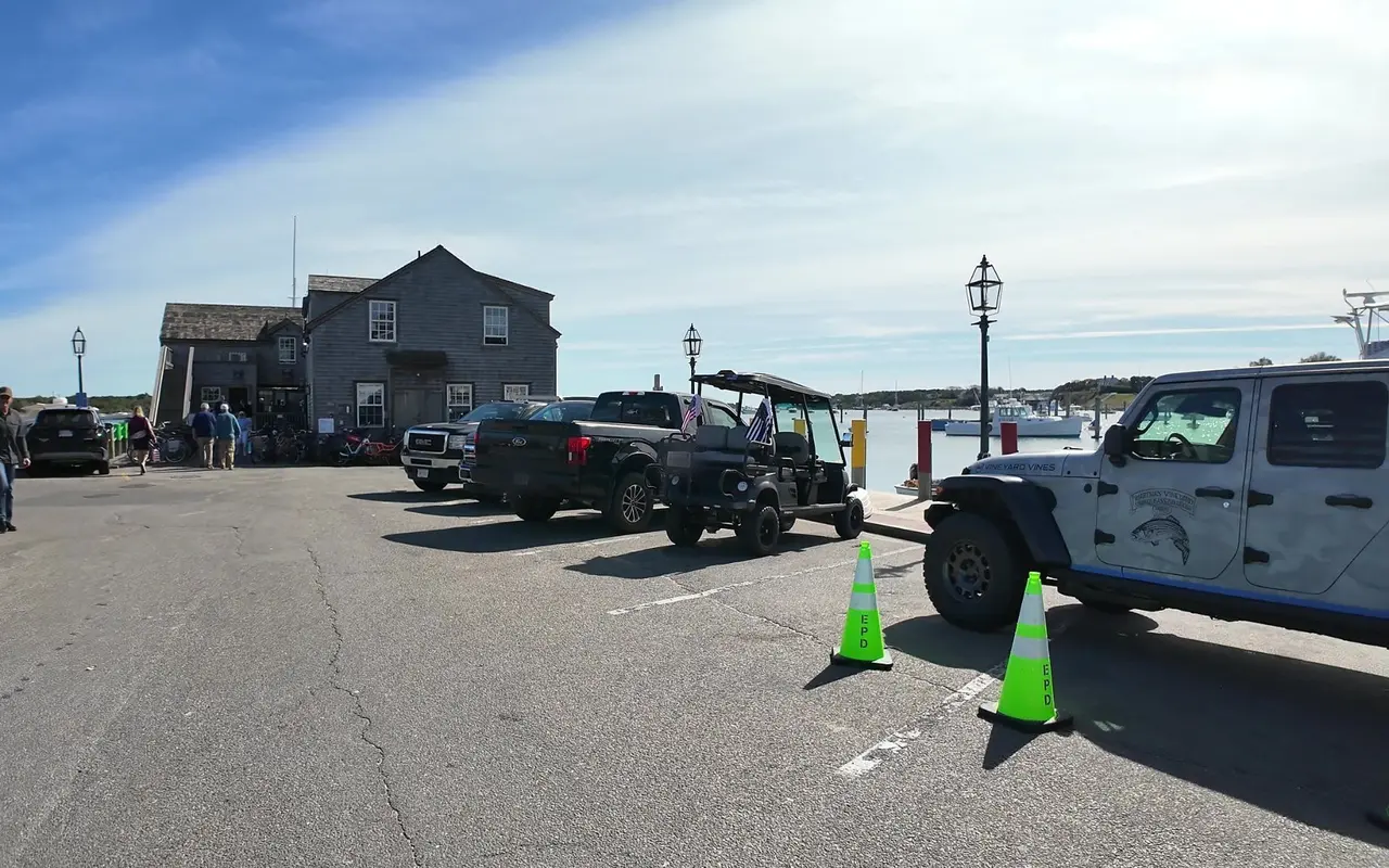 The public parking lot on Dock Street in Edgartown with pickups, a golf cart and parked cars beside the harbor, the shingled Derby headquarters building behind