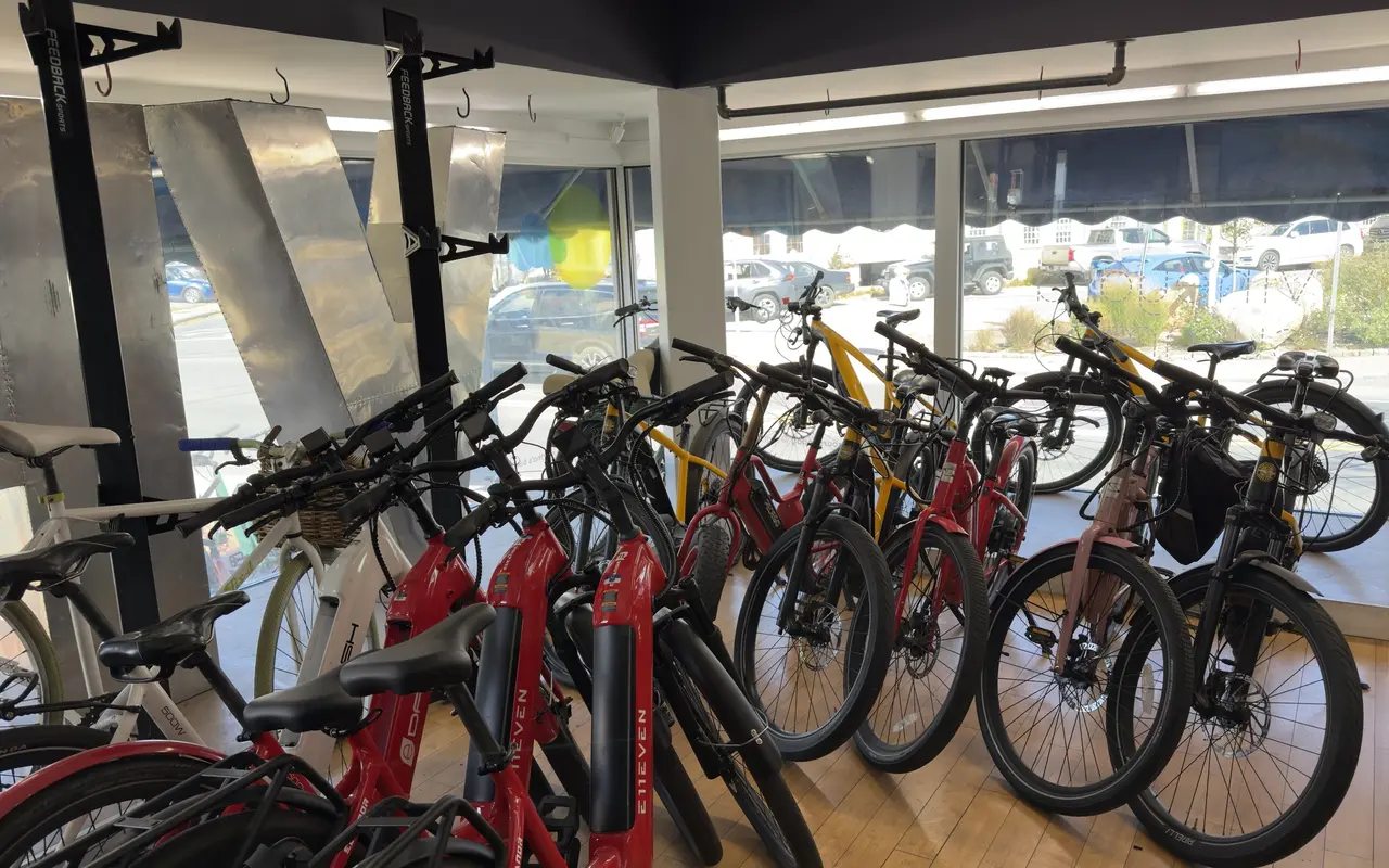 Rows of red, yellow, black and white rental e-bikes inside a bike shop in Vineyard Haven, parked cars visible through the windows