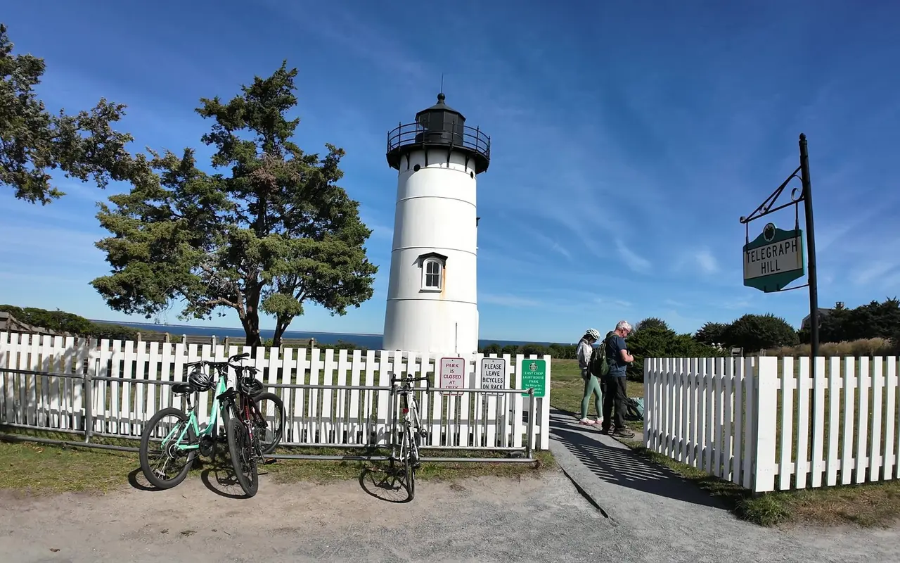 East Chop Lighthouse behind a white picket fence on Telegraph Hill, bicycles at a rack by the gate, signs reading Park Is Closed at Dusk and Please Leave Bicycles on Rack, and a Telegraph Hill sign on a post
