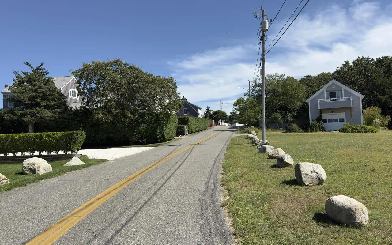 A quiet two-lane road on East Chop in Oak Bluffs under a blue September sky: a row of boulders along a mown verge, a grey-shingled cottage with a balcony over the garage on the right, big shingled houses behind tall hedges on the left
