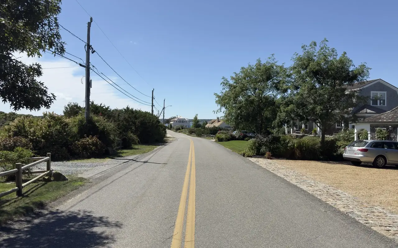 Sunny two-lane road, a split-rail fence on the left, a grey-shingled house and cobblestone-edged drive on the right