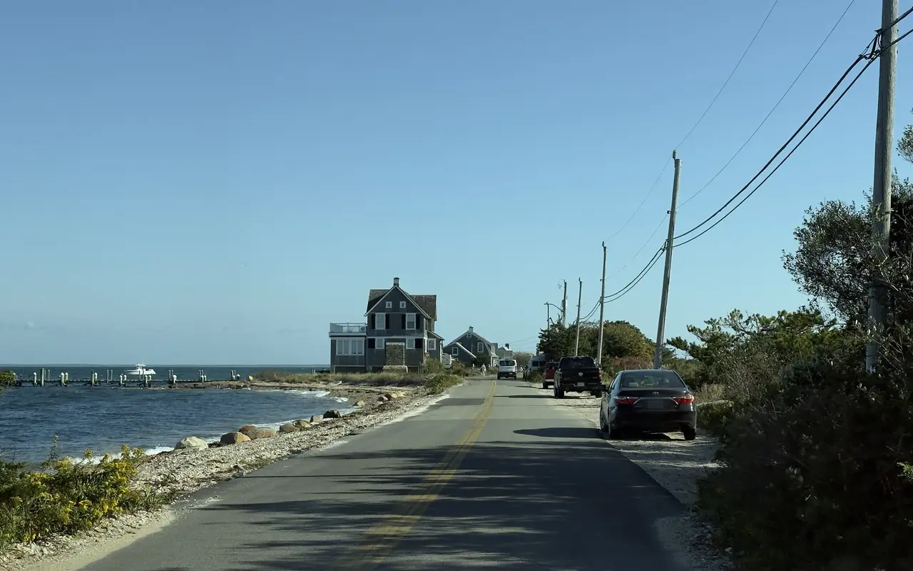 Two-lane road running beside a pebble shore and boulders, a large shingled house and a long dock on the water ahead, parked cars on the right