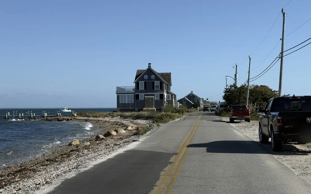 Narrow road beside a pebble shore with pickups and cars parked on the sandy shoulder, a big shingled house and a long dock ahead