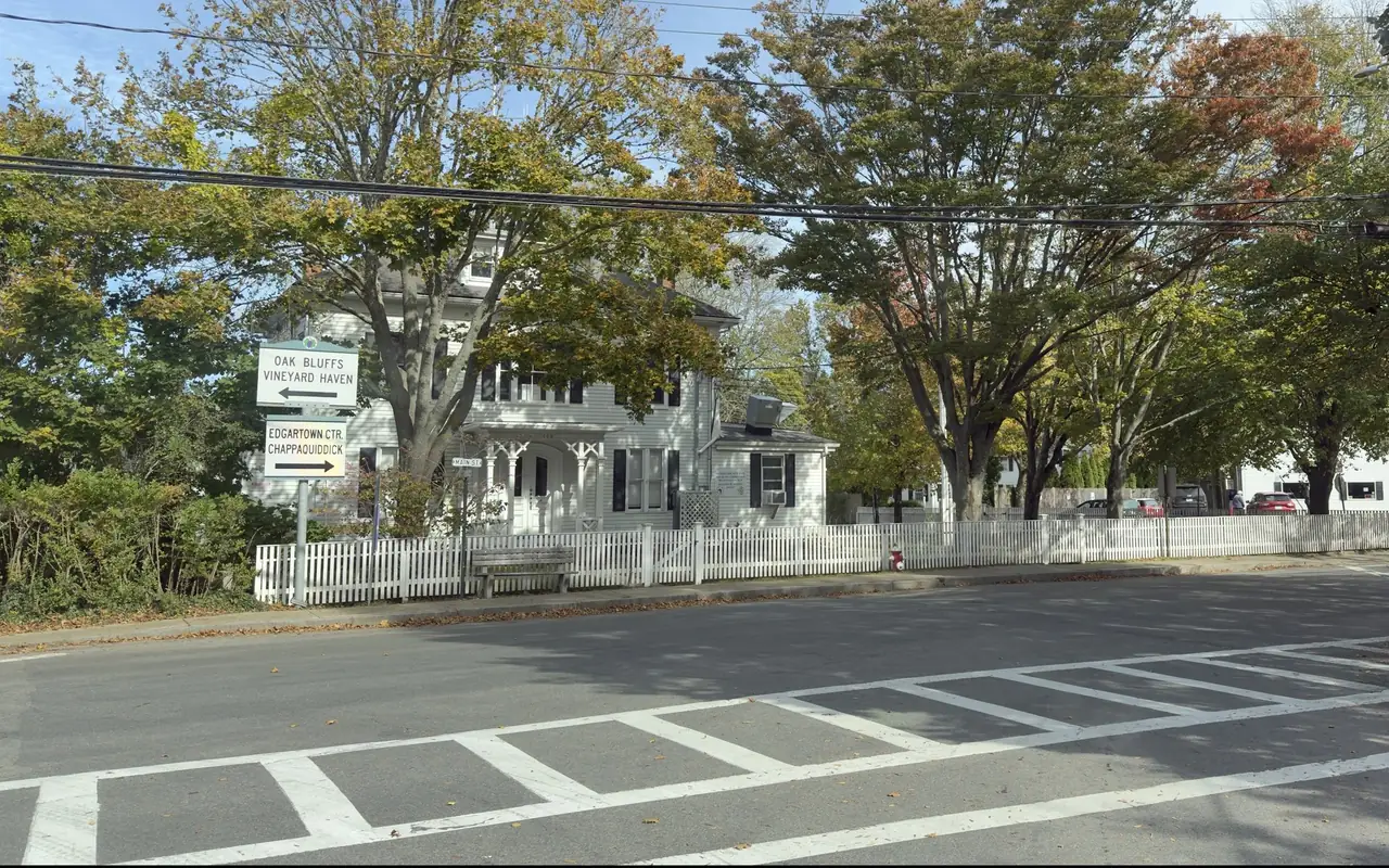A white clapboard building with black shutters behind a white picket fence on Main Street in Edgartown, early fall trees above, direction signs for Oak Bluffs, Vineyard Haven, Edgartown center and Chappaquiddick, and a crosswalk in front