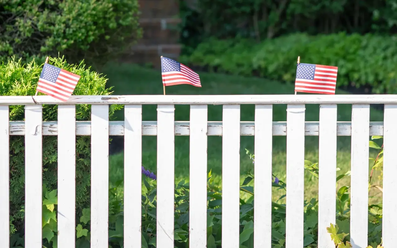 White picket fence decorated with small American flags under green trees