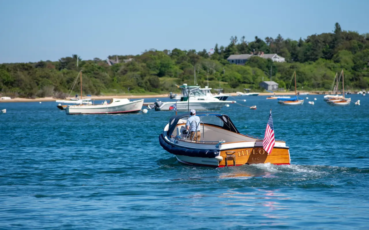 A varnished wooden runabout with an American flag crossing blue Edgartown harbor among moored boats, a green shore with houses behind