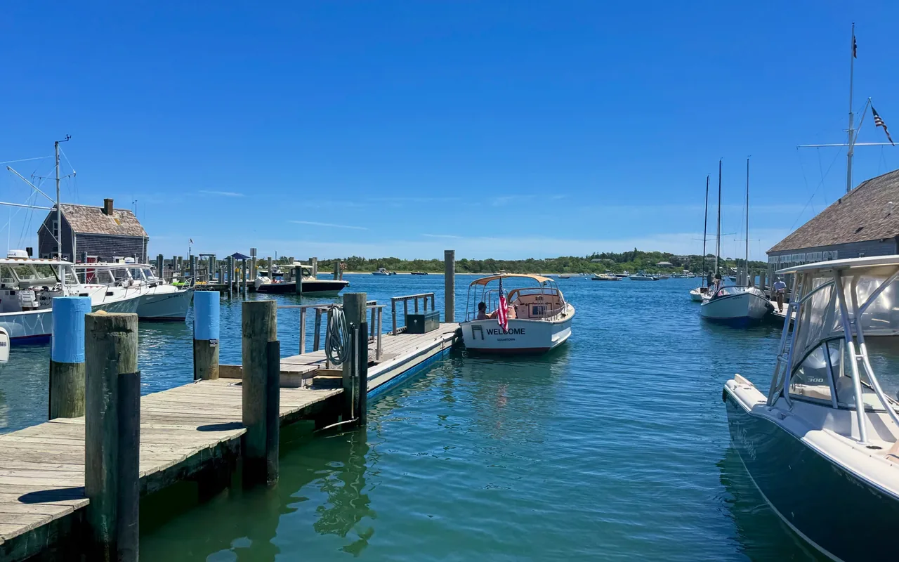 Wooden docks and pilings on Edgartown Harbor under a clear blue sky, a small boat with an American flag and the word Welcome on its stern, sailboats and powerboats moored, the green Chappaquiddick shore across the water
