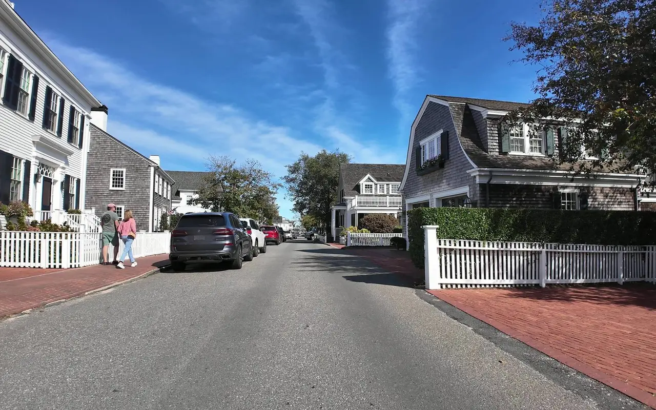 A quiet Edgartown street of white clapboard and gray-shingled houses with brick sidewalks, white picket fences and parked cars under a blue sky