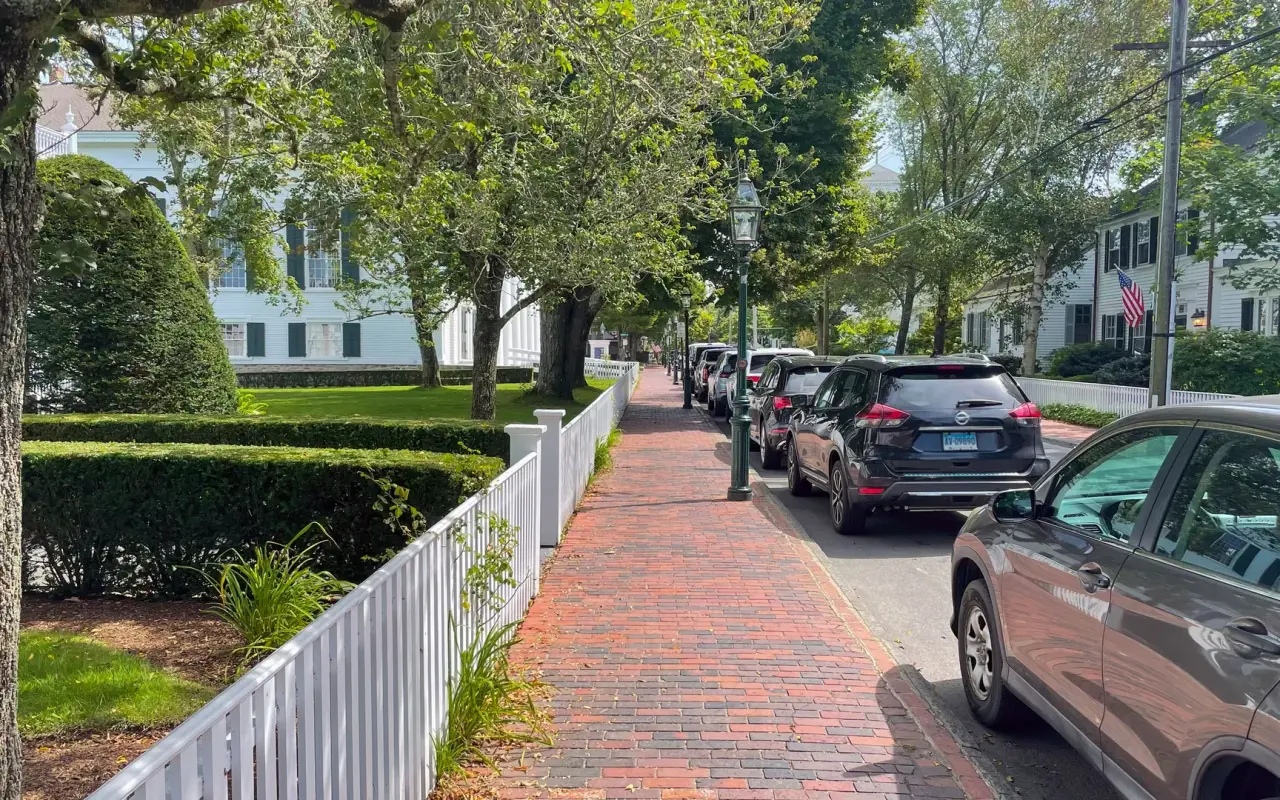 A brick sidewalk along a white picket fence and parked cars on a tree-lined Main Street in Edgartown