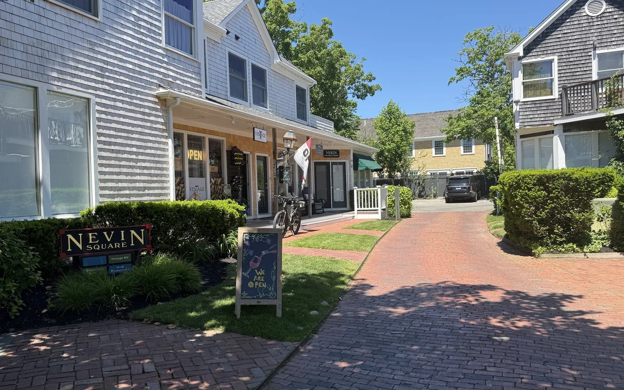 A brick-paved courtyard in Edgartown with a Nevin Square sign, gray-shingled shops with a porch, an Open sign and a chalkboard reading We Are Open, a bicycle parked by the door and green hedges