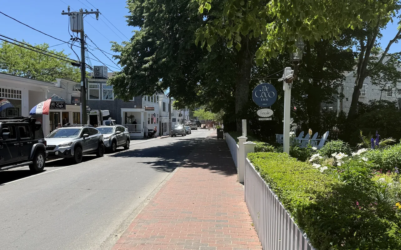 A shady brick sidewalk along North Water Street in Edgartown beside a white picket fence and a flower garden, a round blue sign for The Coco Inn and a l’etoile sign on a white post, parked cars and shopfronts across the street