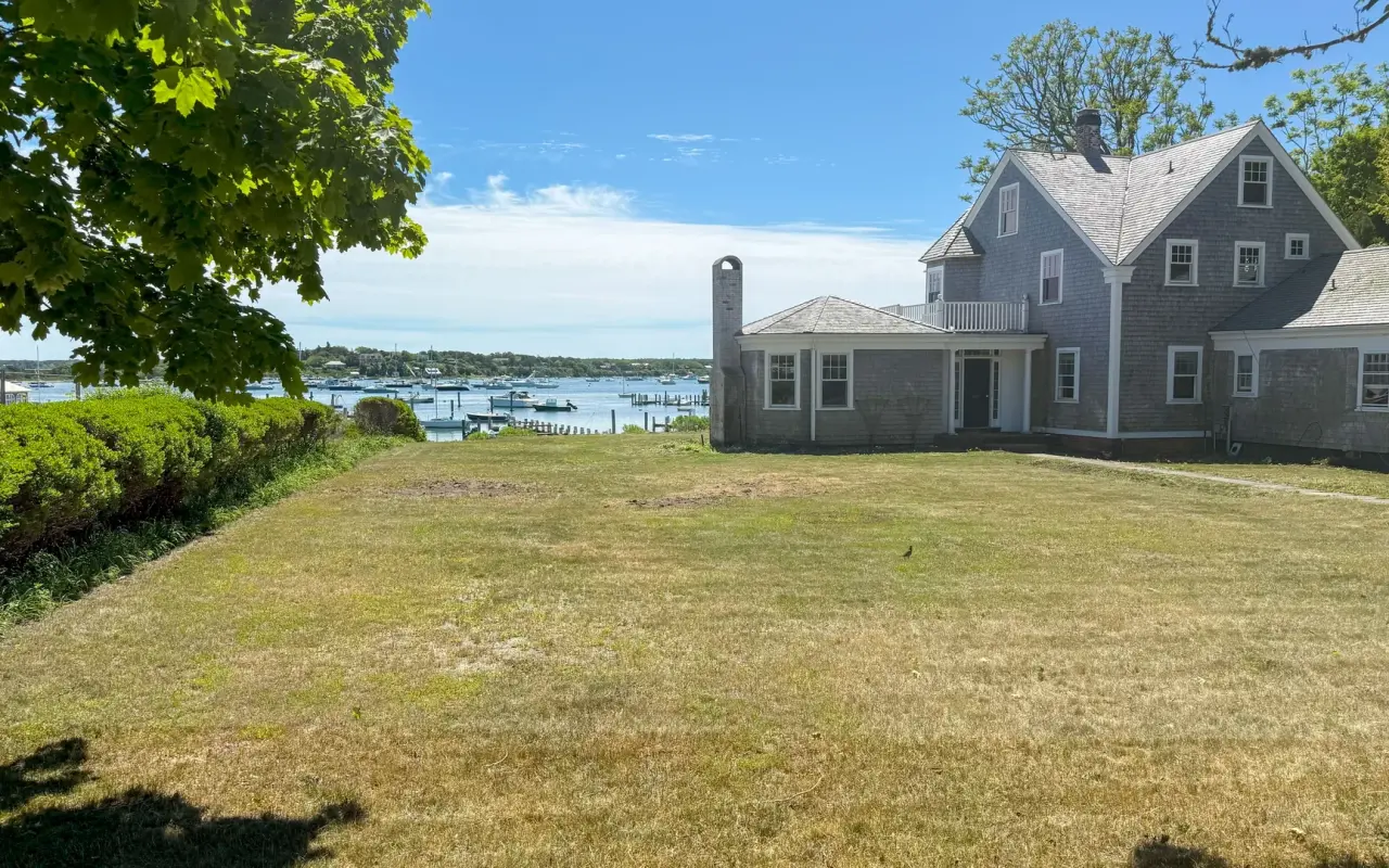 Grassy lawn beside a gray shingled house sloping toward Edgartown harbor with boats in the distance