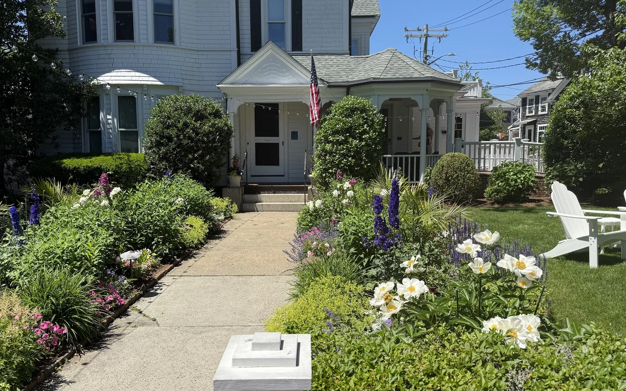 A white Victorian house with a turret and a front porch flying an American flag, a concrete path between flower beds of peonies, delphiniums and salvia, white Adirondack chairs on a green lawn