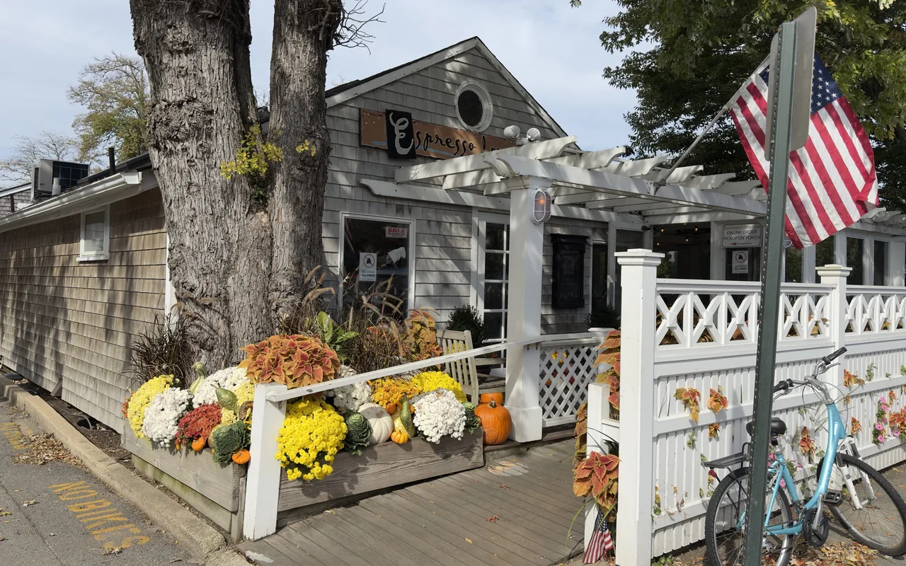 A gray-shingled café with a white pergola and fence, fall mums and pumpkins in a planter box and a blue bicycle leaning on the fence