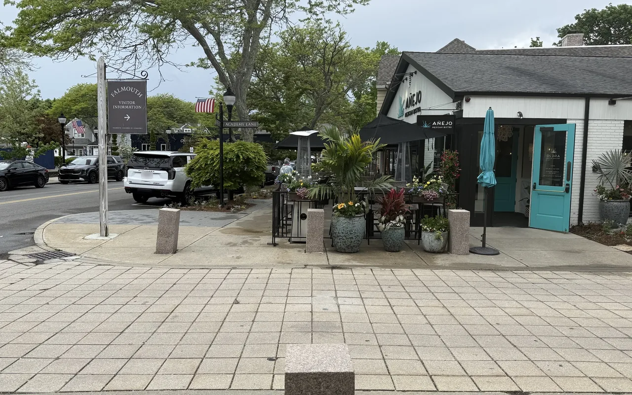 A corner on Main Street in Falmouth with a hanging Falmouth Visitor Information sign, an Academy Lane street sign, and the white building of Anejo Mexican Bistro with a turquoise door and a patio full of potted plants