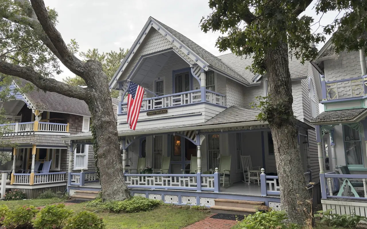 A white gingerbread cottage with lavender trim in the Oak Bluffs Campground, an American flag on the balcony, a Family Treasure name board and rocking chairs on the porch