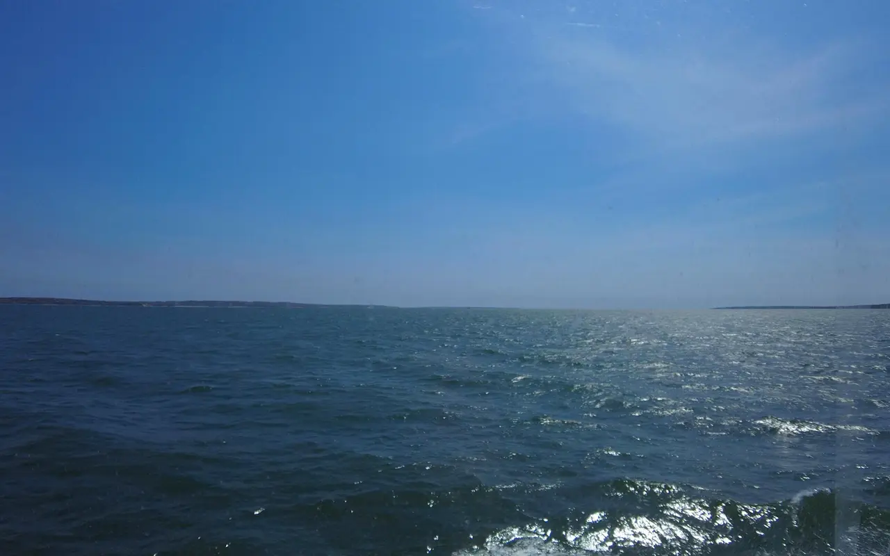 Open water and a distant low shoreline seen through a ferry window on a sunny day, with spots on the glass