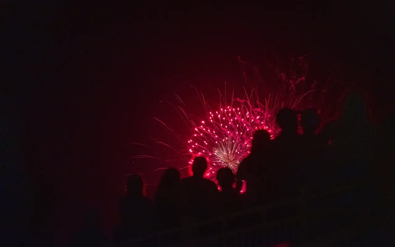 Silhouetted spectators along the Edgartown waterfront lit pink by a firework bursting low over the harbor