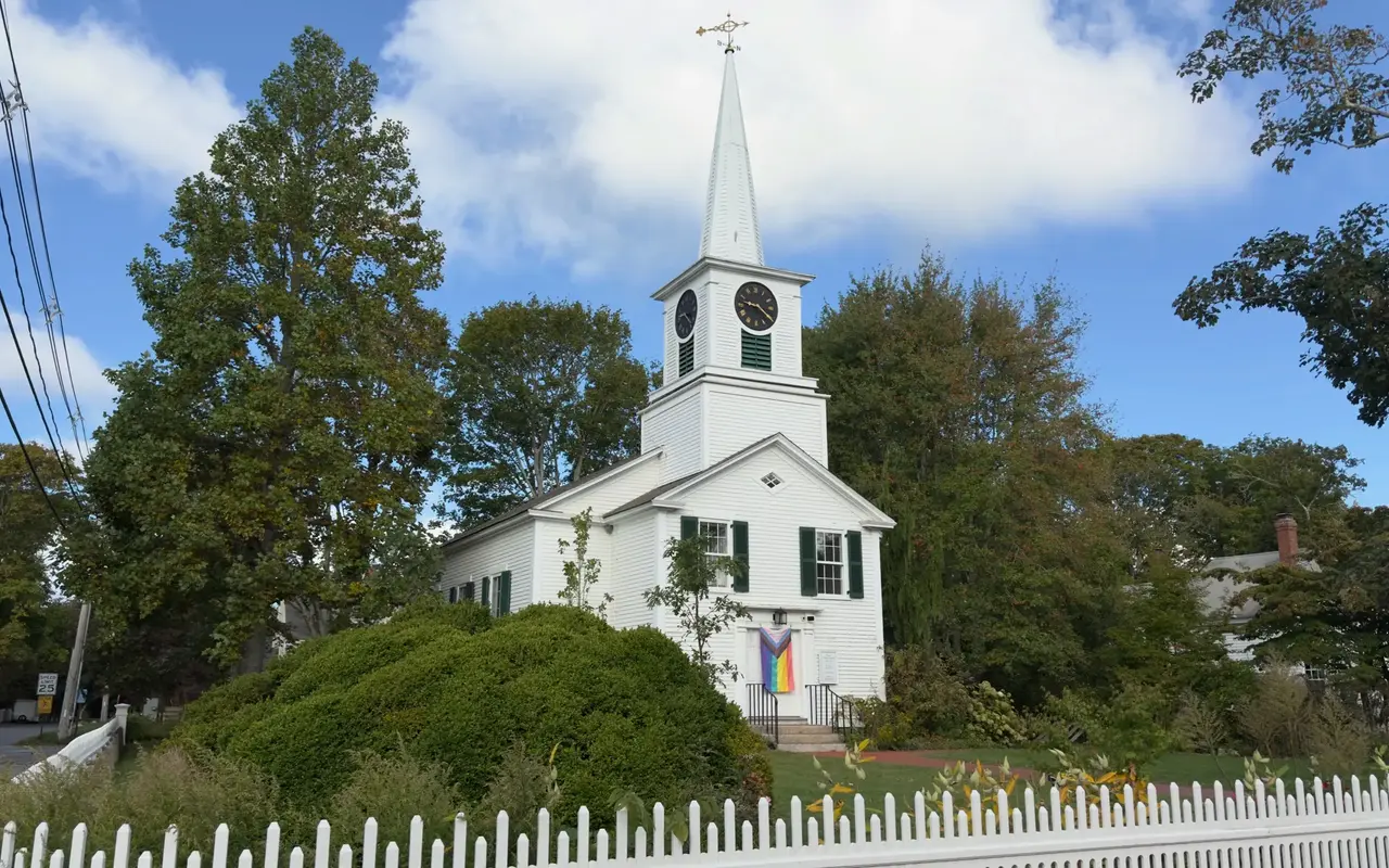 White First Congregational Church of West Tisbury with a clock steeple, green shutters and a Pride flag on the door, behind a picket fence