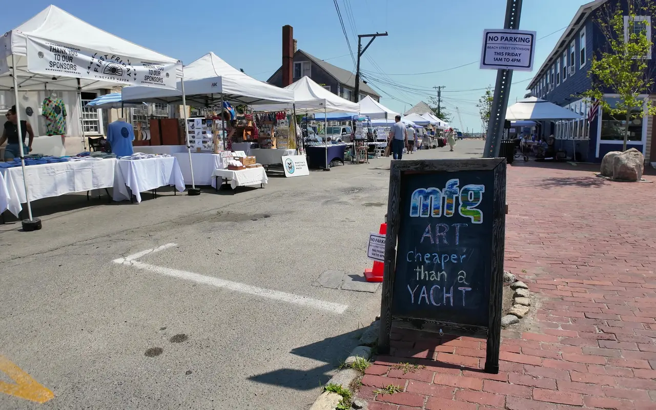 Chalkboard sign reading Art cheaper than a yacht on a brick sidewalk, white vendor tents along Beach Street Extension behind it