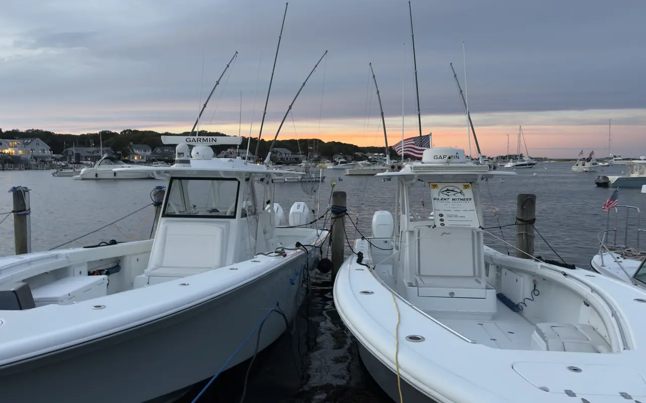 Two white center-console fishing boats side by side in Oak Bluffs harbor at sunset, rods and outriggers pointing at an orange and grey sky, a large flag on the far shore, houses and moored boats behind