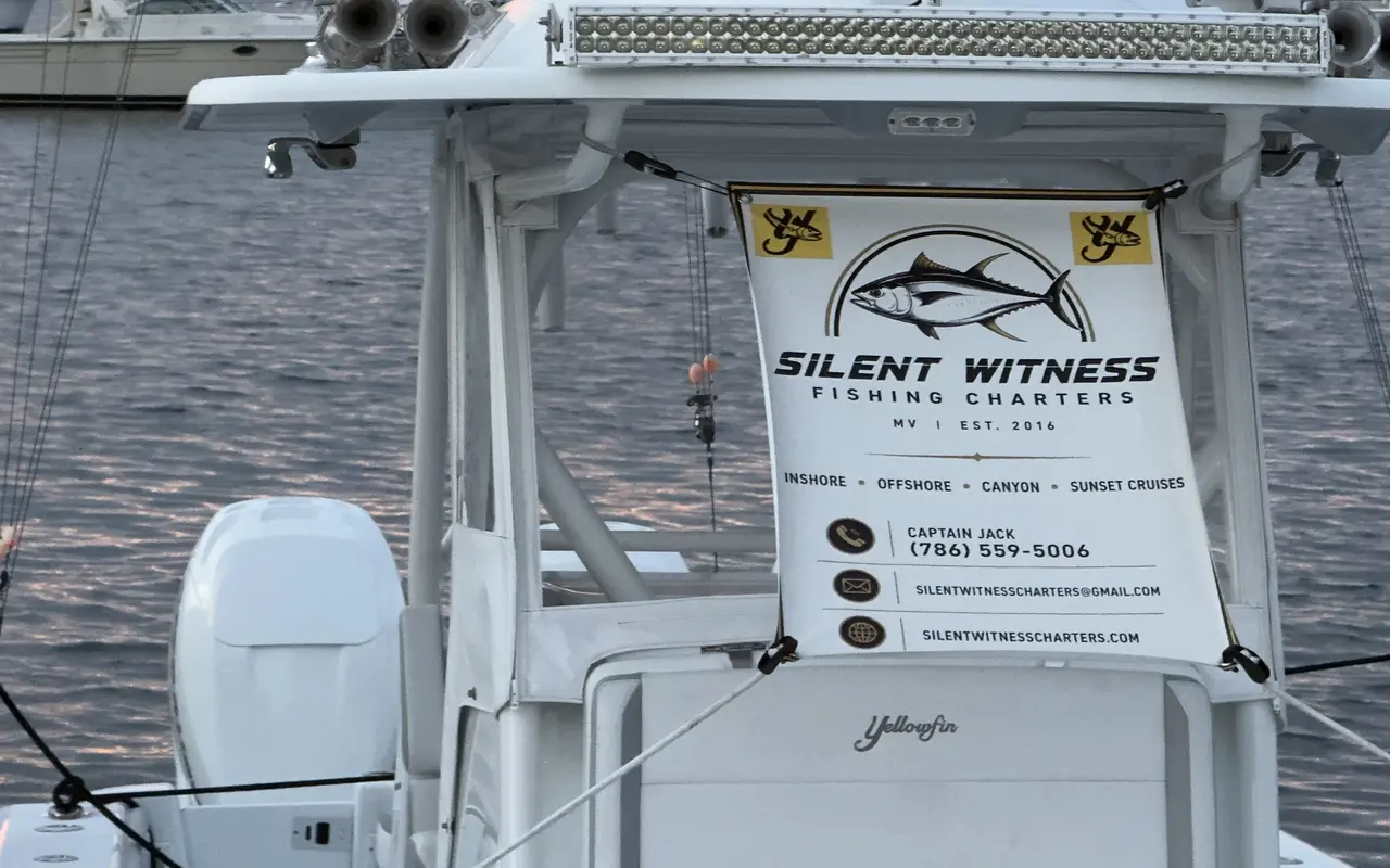 A charter banner strung under the hardtop of a white Yellowfin center-console in Oak Bluffs harbor: a tuna logo, the words inshore, offshore, canyon and sunset cruises, a light bar and outriggers above, calm water behind