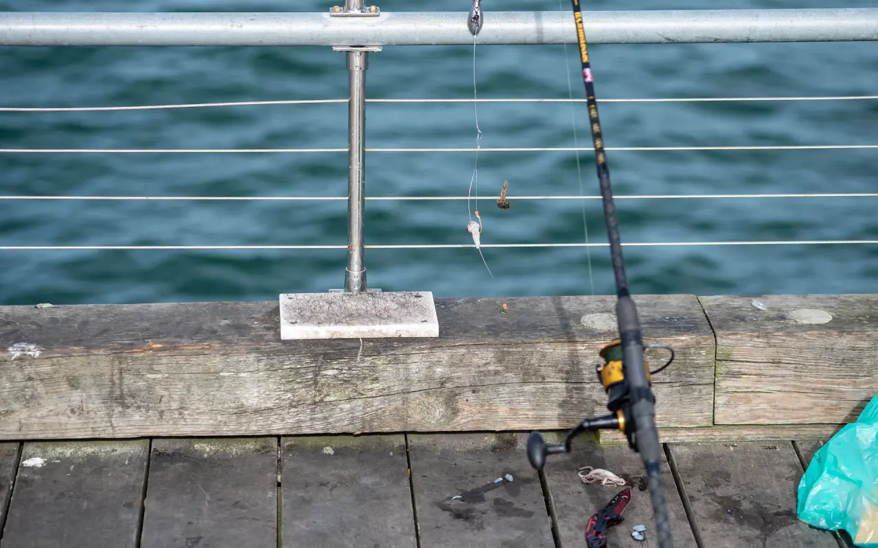 Fishing rod leaning against a pier railing with a baited rig hanging and tackle on the wooden deck
