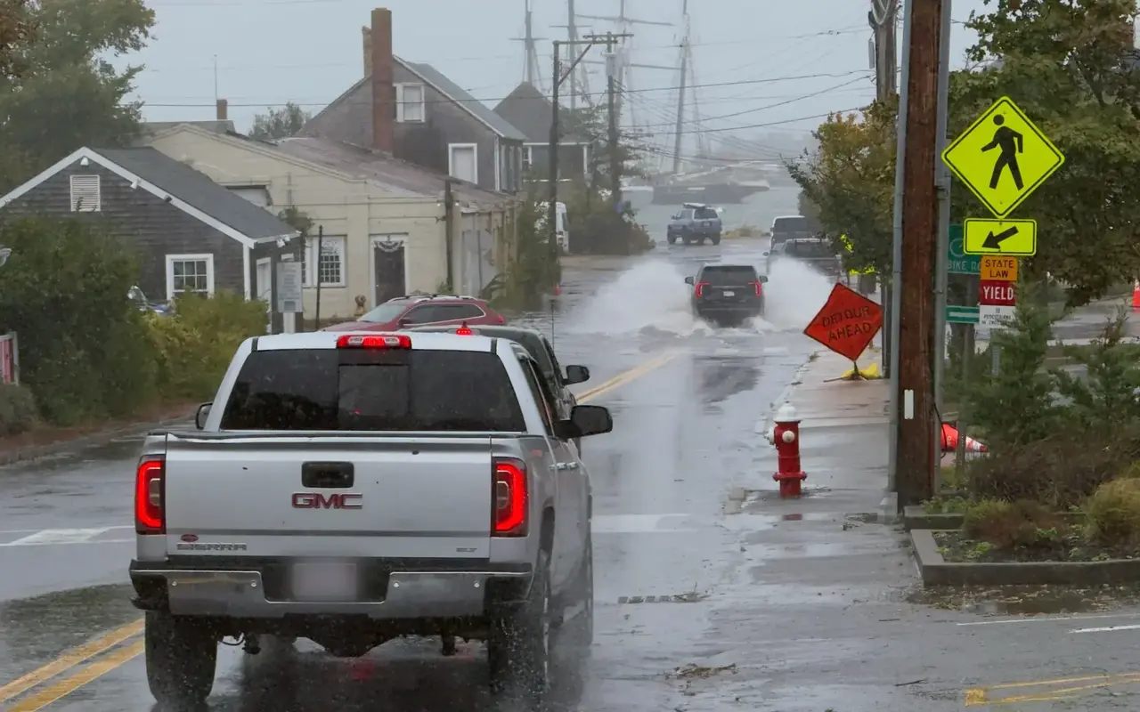 A flooded road at Five Corners in Vineyard Haven during the September 26, 2026 nor'easter: a pickup waiting, an SUV throwing spray as it drives through the water toward the harbor, an orange Detour Ahead sign on the sidewalk, a red hydrant and tall ship masts in the rain behind