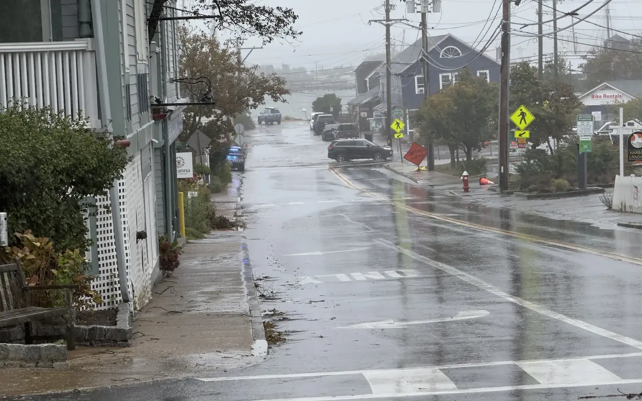 Main Street in Vineyard Haven seen from beside the Mansion House during the September 26, 2026 nor'easter: a wet street running downhill to Five Corners, a police cruiser with blue lights, an orange Detour Ahead sign, a red hydrant and floodwater across the bottom of the street, harbor masts in the mist