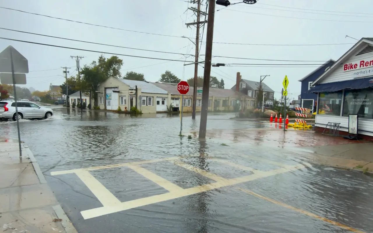 Five Corners in Vineyard Haven flooded during a nor'easter on September 26, 2026: water across the whole intersection, a silver SUV wading through, orange cones, barricades and yellow sandbags outside Martha's Bike rental shop, a stop sign and utility poles standing in the water under a grey sky