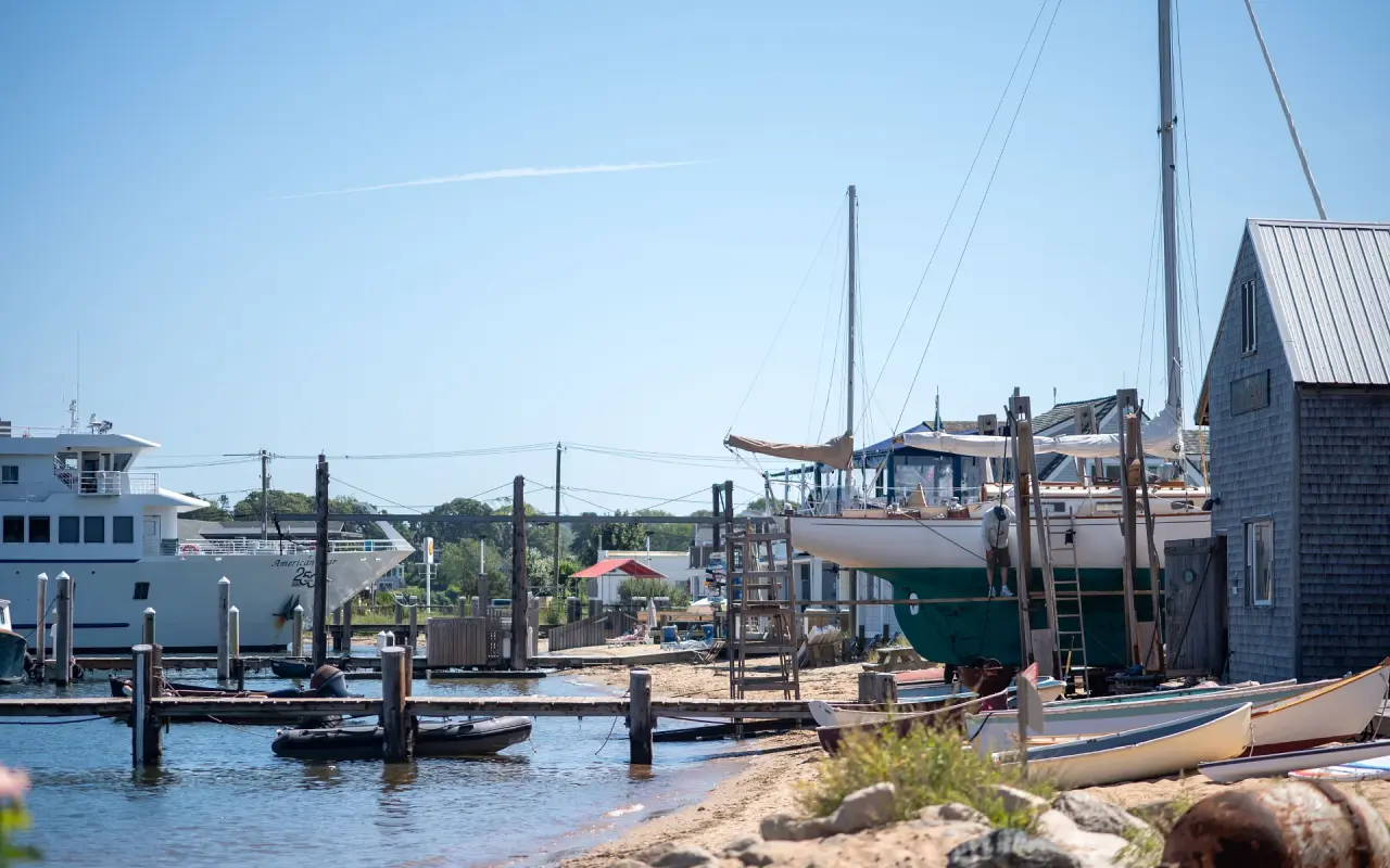 Boatyard on a harbor beach with a wooden sailboat hauled out, small skiffs on the sand and a ship in the background