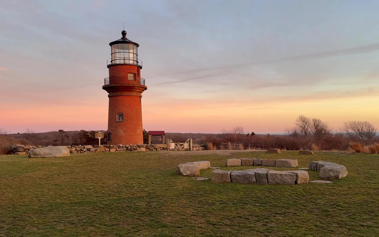 Red brick Gay Head Light at dusk under a pink and blue sky, a green lawn and a ring of low stones in front