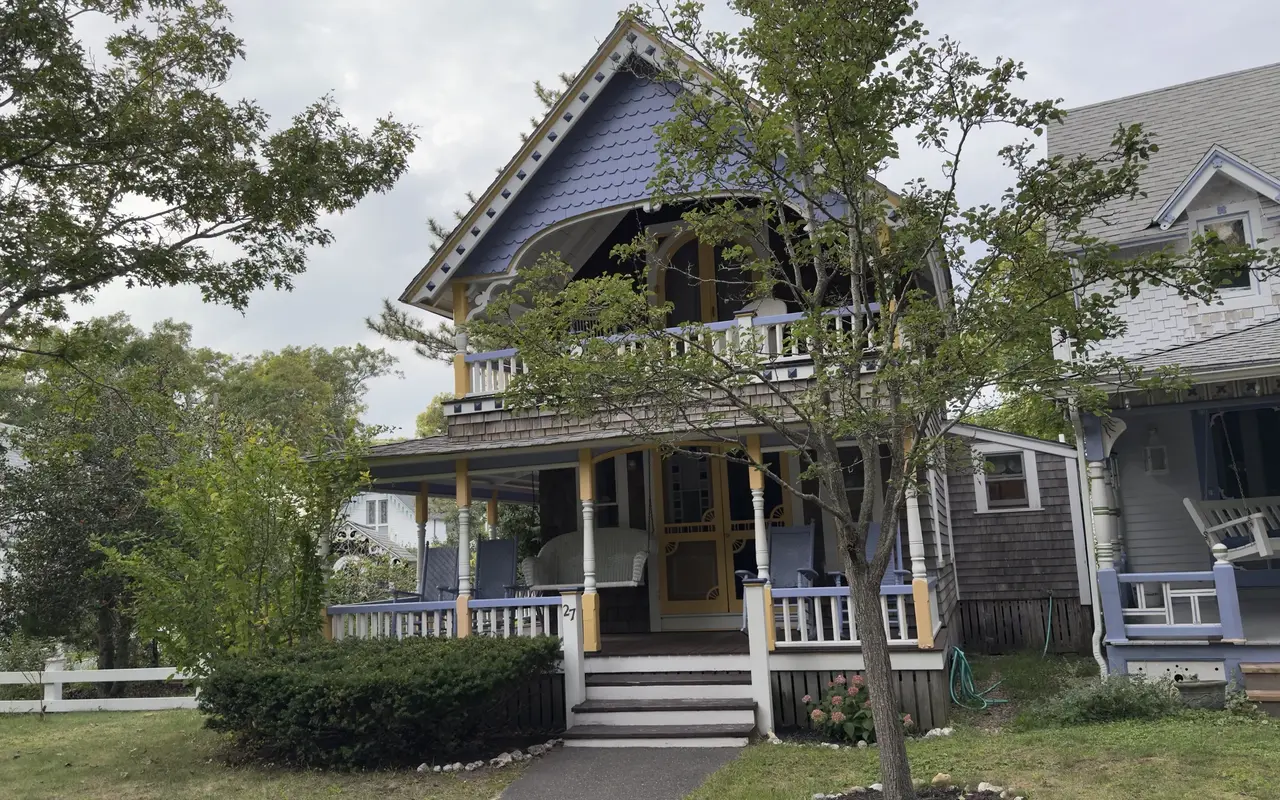A gingerbread cottage in the Oak Bluffs Campground with a purple fish-scale gable, yellow posts, a white balcony and blue chairs on the porch, number 27