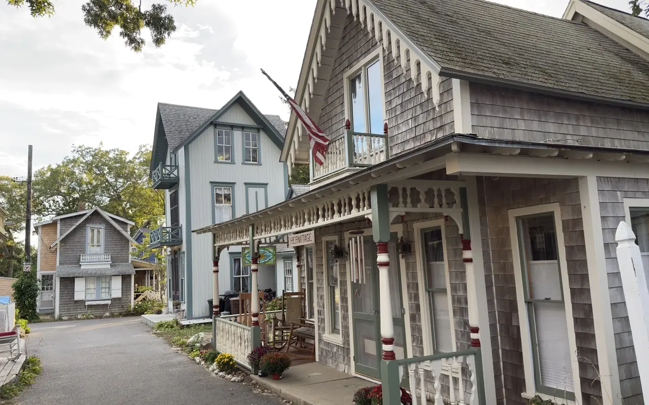 A gray-shingled cottage named Hermitage in the Oak Bluffs Campground with cream gingerbread trim, a stained-glass panel on the porch, an American flag and a lane of cottages beyond