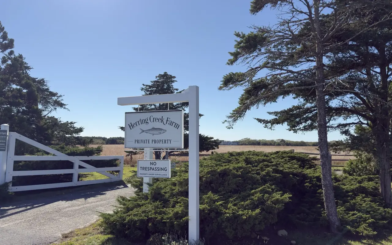 White farm gate and hanging sign reading Herring Creek Farm, Private Property, with a No Trespassing sign below and a No Beach Access sign on the gate post, pines and open tan fields behind under a clear sky