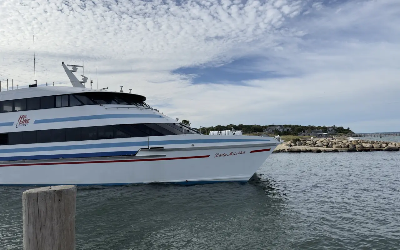 The Hy-Line Cruises fast ferry Lady Martha, white with blue stripes, turning past the stone jetty into Oak Bluffs harbor under a mackerel sky, a wooden piling in the foreground and the town pier in the distance