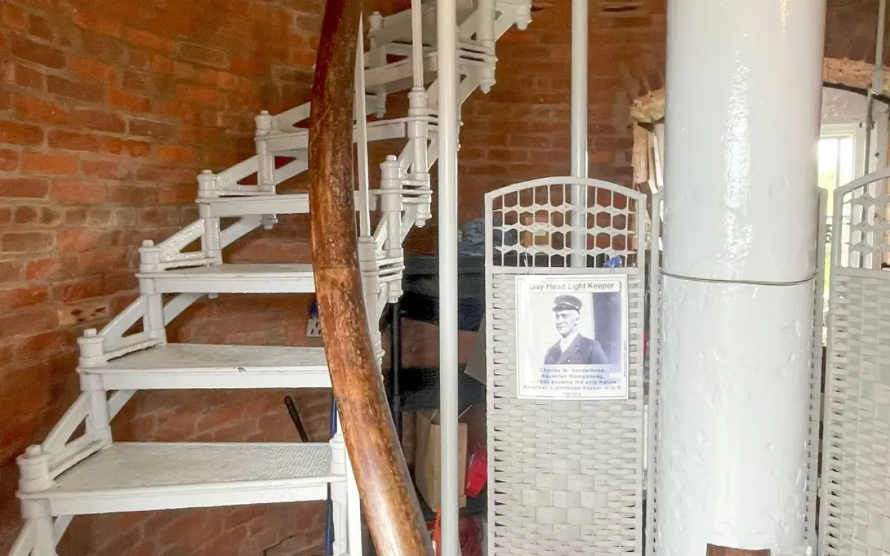 A white spiral staircase inside the red brick Gay Head Lighthouse with a framed portrait on the wall
