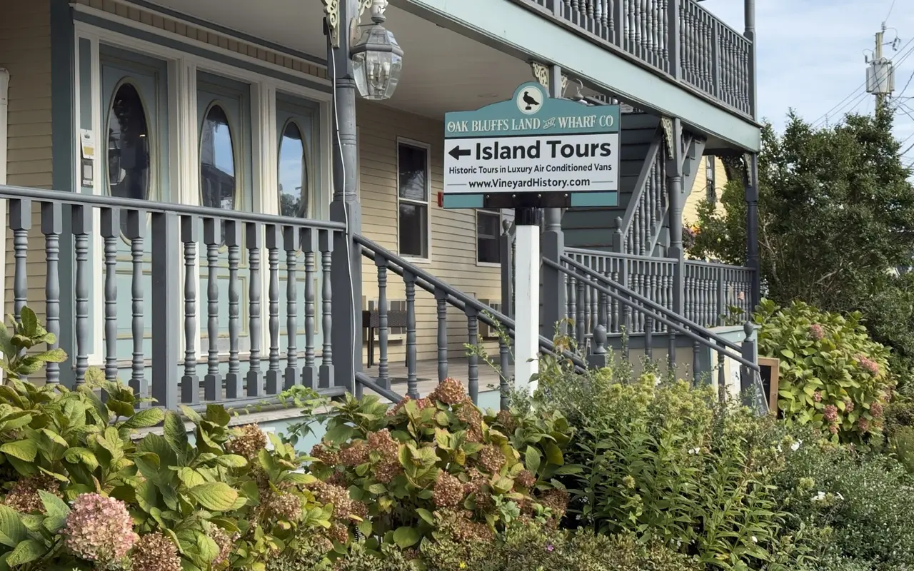 A sign for the Oak Bluffs Land and Wharf Co pointing to Island Tours, historic tours in air-conditioned vans, in front of a yellow Victorian building with grey porch railings and a double balcony, hydrangeas going pink below