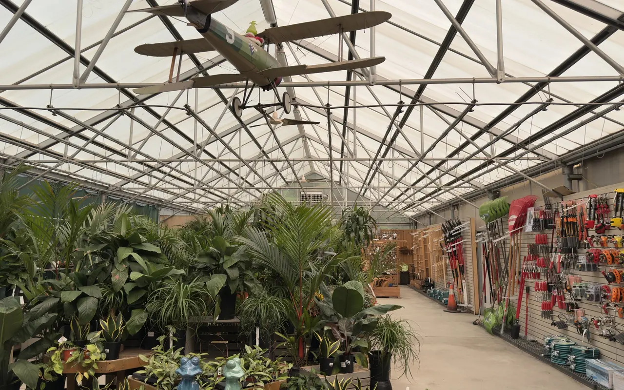 Greenhouse interior at Jardin Mahoney garden center in Oak Bluffs, with tropical houseplants on one side, a wall of hand tools and hoses on the other, and a model biplane hanging from the roof trusses
