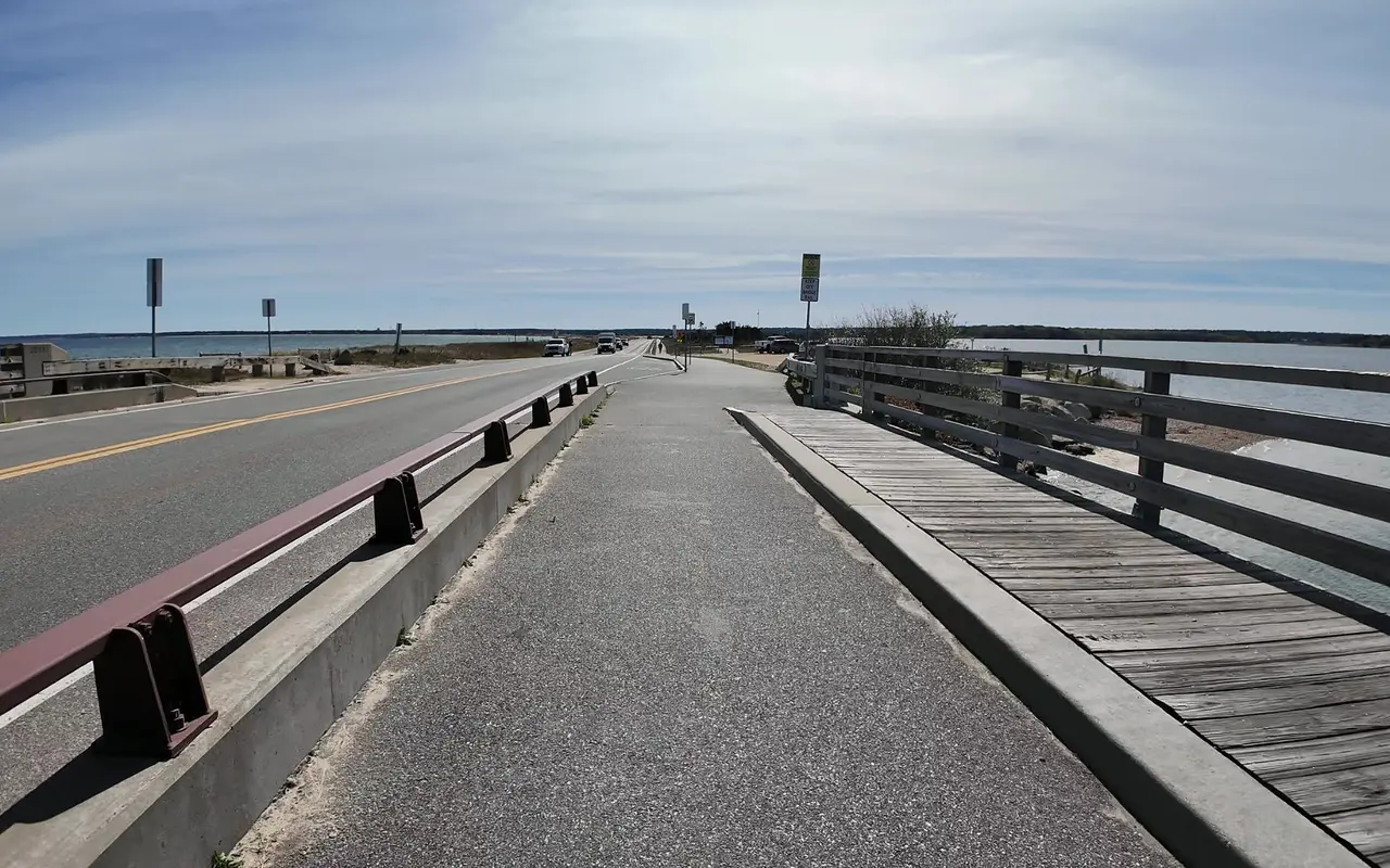 The bike path over the Jaws bridge seen from a bicycle, the road on the left, a wooden rail on the right with a No Jumping Shallow Water sign and a Keep Off Bridge Rail sign ahead