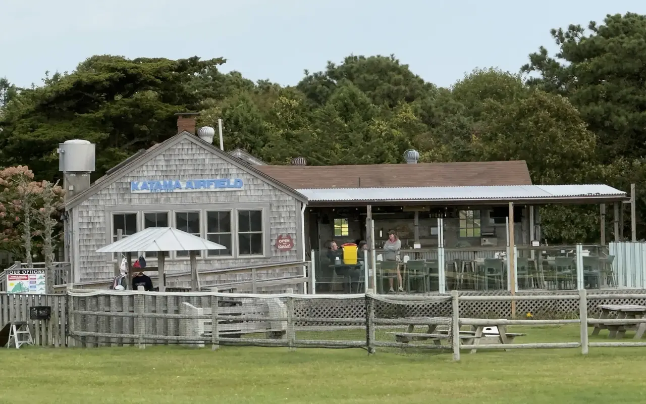 Shingled Katama Airfield building with a covered deck behind glass panels, picnic tables on the lawn and a split-rail fence in front