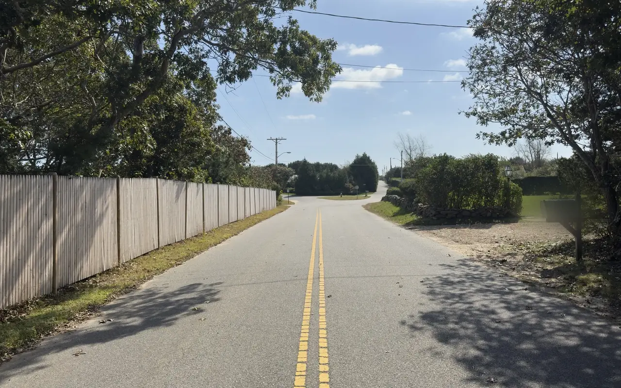 Edgartown Bay Road in Katama with a tall wooden stockade fence on the left, a mailbox at right and a small grassy island at the junction ahead