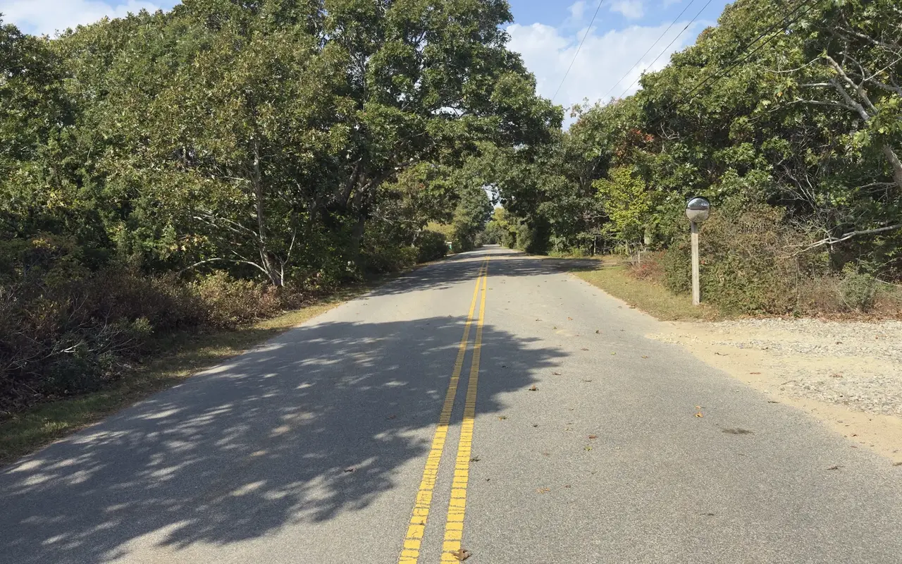 Empty Edgartown Bay Road in Katama with a double yellow line running between oak trees and scrub, a round traffic mirror on a post at right