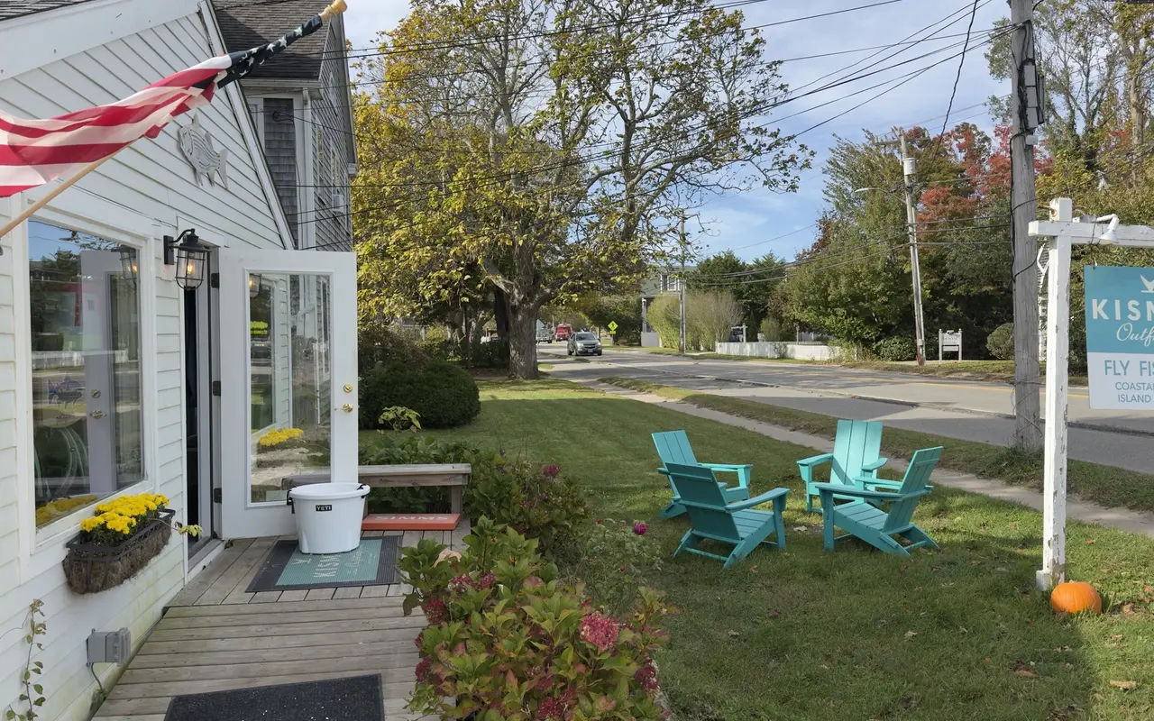 Porch of Kismet Outfitters in Edgartown with an American flag, a white bucket on the step and four blue Adirondack chairs on the lawn beside the road