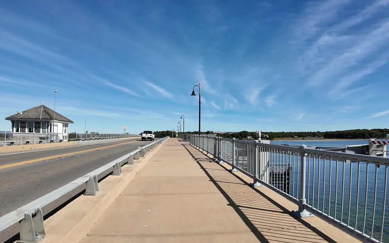 The wide sidewalk across the Lagoon Pond drawbridge with a gray railing and lamp posts, the roadway on the left and a small shingled bridge tender house at the far end, under a blue sky