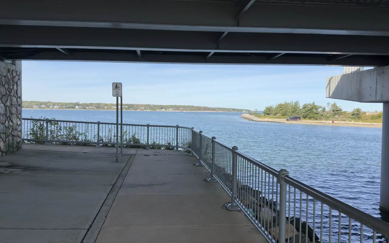 Concrete walkway with a metal railing under the Lagoon Pond drawbridge, a Walk Your Bike sign, the channel and a wooded point beyond