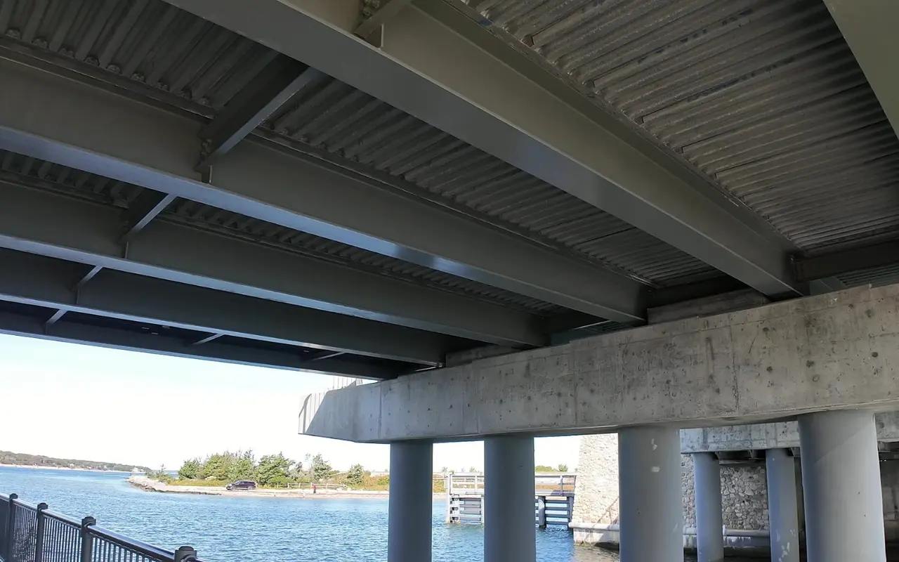 The underside of the Lagoon Pond drawbridge with steel girders and round concrete piers, a railed walkway on the left and the channel and a wooded point beyond