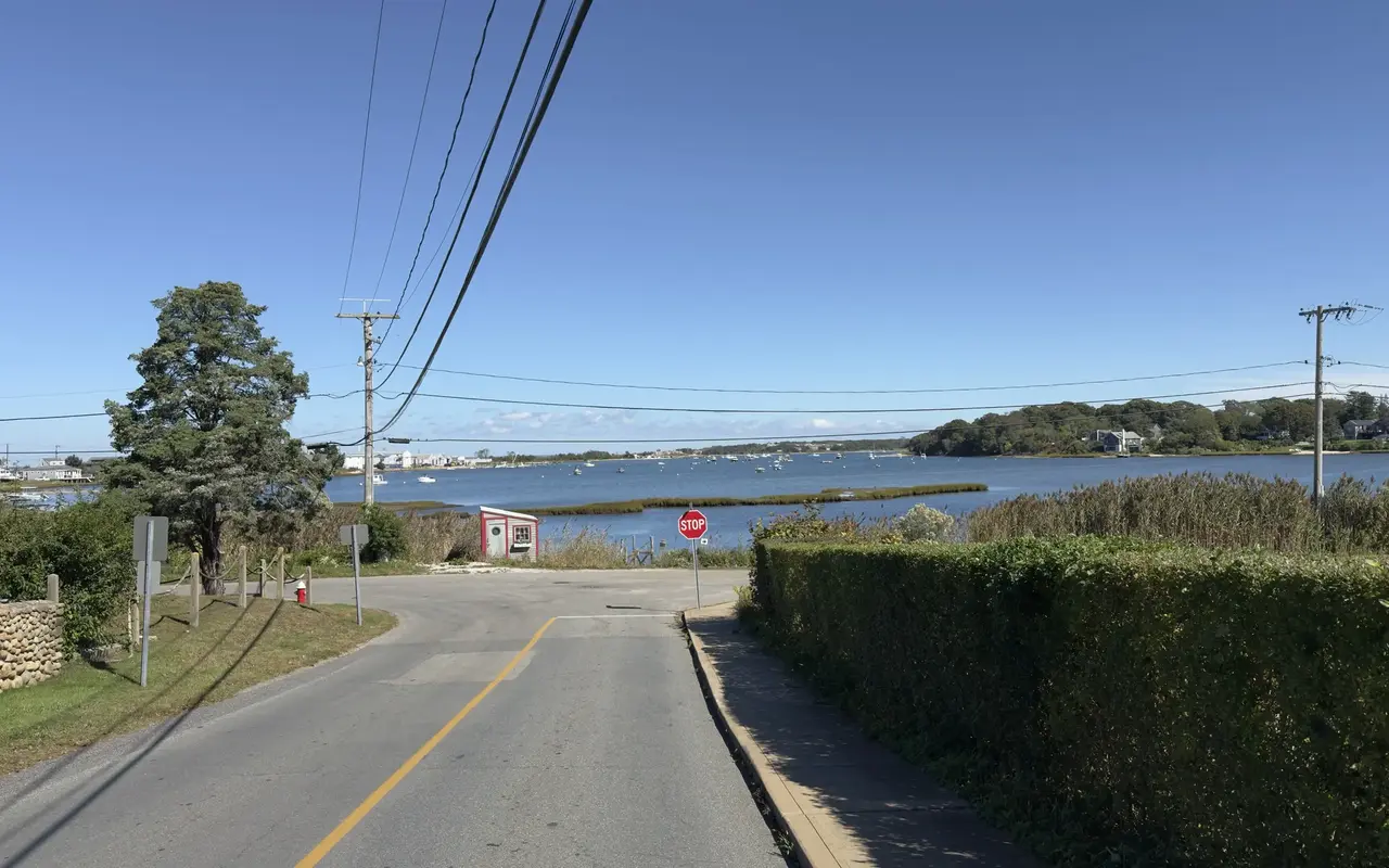 A quiet road leading down to Lagoon Pond with moored boats and a marsh under a blue sky