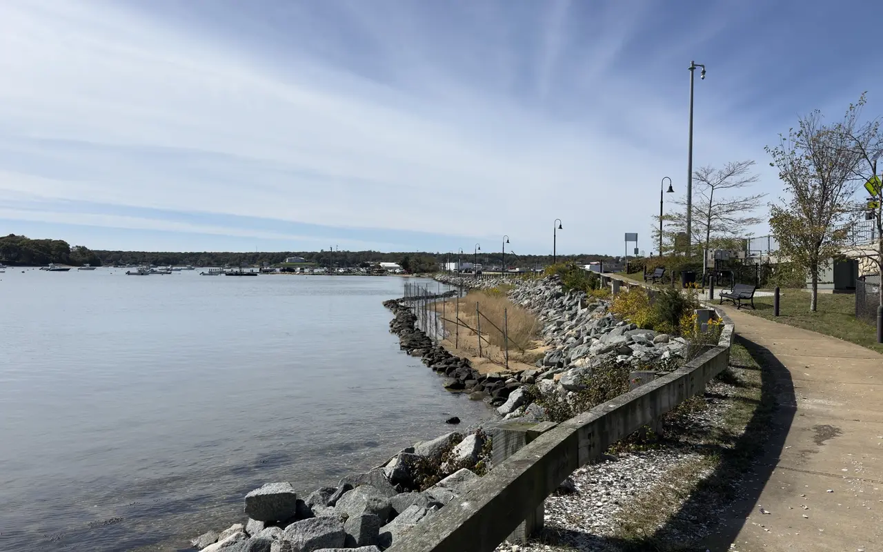 A paved path with benches and lamp posts beside a rocky shoreline and calm water, boats moored in the distance, on the Vineyard Haven side of the Lagoon Pond bridge